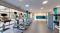Located at 2701 Neabsco Common Pl in Woodbridge, VA, this well-appointed fitness center offers a generously sized, single-floor exercise facility designed to support a full range of workout routines. The space is thoughtfully divided into distinct functional zones, each catering to different training needs.

The cardio area lines the perimeter near expansive floor-to-ceiling windows, allowing abundant natural light to fill the room while offering views of the outdoors. Treadmills, ellipticals, and upright cycling machines are neatly arranged in this section. A striking teal accent wall and matching teal-wrapped structural columns provide a bold, energizing aesthetic throughout the space.

The strength and free weights area features a dumbbell rack, weight benches, cable machines, and cable resistance systems, all positioned on durable, speckled rubber flooring designed for high-impact use. Wall-mounted mirrors in this section enhance the sense of space and allow users to monitor their form.

A dedicated hardwood studio floor anchors a separate zone ideal for stretching, yoga, group fitness, or functional training. Medicine balls and other accessories are neatly stored nearby. Glass-paneled walls and doors border this area, maintaining an open, airy feel throughout.

The facility features a drop ceiling with recessed and flush lighting panels, ensuring consistent illumination across all areas. The overall design is modern, clean, and functional, creating an inviting environment for fitness enthusiasts of all levels.