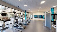 Located at 2701 Neabsco Common Pl in Woodbridge, VA, this well-appointed fitness center offers a generously sized, single-floor exercise facility designed to support a full range of workout routines. The space is thoughtfully divided into distinct functional zones, each catering to different training needs.

The cardio area lines the perimeter near expansive floor-to-ceiling windows, allowing abundant natural light to fill the room while offering views of the outdoors. Treadmills, ellipticals, and upright cycling machines are neatly arranged in this section. A striking teal accent wall and matching teal-wrapped structural columns provide a bold, energizing aesthetic throughout the space.

The strength and free weights area features a dumbbell rack, weight benches, cable machines, and cable resistance systems, all positioned on durable, speckled rubber flooring designed for high-impact use. Wall-mounted mirrors in this section enhance the sense of space and allow users to monitor their form.

A dedicated hardwood studio floor anchors a separate zone ideal for stretching, yoga, group fitness, or functional training. Medicine balls and other accessories are neatly stored nearby. Glass-paneled walls and doors border this area, maintaining an open, airy feel throughout.

The facility features a drop ceiling with recessed and flush lighting panels, ensuring consistent illumination across all areas. The overall design is modern, clean, and functional, creating an inviting environment for fitness enthusiasts of all levels.