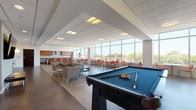 The Innova Game Room in Cleveland, Ohio is a thoughtfully designed, single-floor recreational and social space that combines entertainment, comfort, and functionality in a bright, contemporary setting. Floor-to-ceiling windows line the perimeter, flooding the interior with abundant natural light and offering sweeping views of the Cleveland skyline and surrounding treetops, creating an open and airy atmosphere throughout.

The main gathering area features a full-sized pool table as a central focal point, complemented by generous lounge seating with modern sofas and armchairs arranged for both social interaction and relaxation. A dedicated media lounge area includes a wall-mounted flat-screen TV flanked by lush indoor plants, with comfortable seating ideal for casual viewing or unwinding.

Along the window-facing wall, high-top café-style tables paired with vibrant coral-red chairs provide additional seating for dining or informal meetings, taking full advantage of the panoramic outdoor views.

The well-appointed kitchen is a standout feature, showcasing warm wood cabinetry, dark granite countertops, stainless steel appliances including dual ranges, a stainless sink, and a subway tile backsplash. A kitchen island with bar-height seating extends the space's social functionality.

Polished concrete and hardwood-style flooring flow throughout, tying together the clean, modern aesthetic. Glass-paneled partition walls define transitional zones while maintaining visual openness across the entire space.