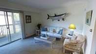 This single-floor residence offers a thoughtfully arranged layout with multiple bedrooms, a central living area, kitchen, and bathroom, all connected by a practical and flowing floor plan. The living room sets a relaxed coastal tone, featuring soft carpeted flooring, light-colored walls, and rattan furniture with blue accent cushions that evoke a beachy, laid-back atmosphere. A striking large fish mount displayed prominently on the wall serves as a standout decorative focal point, reinforcing the nautical theme throughout. Natural light filters into the space through windows, contributing to a bright and airy ambiance. The bedrooms are comfortably sized, each featuring closet storage and carpeted floors, with natural light adding warmth to the interiors. The kitchen is centrally positioned within the home, offering convenient access to the surrounding rooms. A bathroom is also situated near the bedroom wing for easy accessibility. Throughout the residence, the coastal aesthetic is carried consistently, creating a cohesive and inviting character. The overall layout is efficient and well-suited for relaxed living, with each room contributing to the home's warm, casual, and welcoming atmosphere.