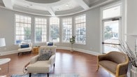 Located at 605 Candler Ln in Charlotte, NC, this single-floor residence showcases refined architectural detailing throughout. The main living area commands attention with its soaring coffered and tray ceilings, which lend a grand sense of verticality and openness to the space. Rich hardwood flooring flows seamlessly underfoot, anchoring the warm, neutral color palette of soft grays and crisp whites that define the interior.

A standout feature of the living room is the floor-to-ceiling stone fireplace, framed by a classic white shiplap surround and substantial mantel, offering a compelling focal point. Flanking the entryway, a built-in bench sits beneath a large arched window, filling the foyer with natural light and creating a welcoming transition into the home.

One of the most distinctive architectural elements is a curved rotunda-style sitting area enveloped by a sweeping arc of tall, grid-paned windows. This light-filled alcove offers expansive outdoor views and is crowned by a circular tray ceiling, making it an exceptional feature for relaxation or entertaining.

The open layout connects the living space to a glimpsed kitchen area beyond a pass-through, reinforcing the home's functional flow. Recessed lighting throughout complements the abundant natural light. Overall, the space combines classical architectural elegance with an airy, sophisticated ambiance.