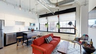 Located at 350 Mount Vernon Ave in Columbus, Ohio, this single-floor loft blends industrial character with modern comfort in a striking open-concept design. Soaring ceilings with exposed ductwork and mechanical systems give the space an authentic urban loft aesthetic, while track lighting and a ceiling fan provide both function and style overhead. Large, multi-pane factory-style windows line the walls, flooding the interior with abundant natural light and offering views of the surrounding Columbus neighborhood. The living area features a spacious open layout anchored by a bold red sofa, a wood coffee table, and a plush area rug, creating a warm and inviting gathering space. The kitchen flows seamlessly into the living area, outfitted with stainless steel appliances including a range and microwave, white subway tile backsplash, dark cabinetry, and a freestanding island with bar stool seating — ideal for casual dining and entertaining. Hanging greenery suspended from the windows adds a lively, organic touch that softens the industrial elements throughout. The overall ambiance strikes a confident balance between raw, warehouse-inspired architecture and thoughtfully curated modern furnishings, making this a distinctive and functional urban living space.