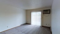 This single-floor residential space offers a straightforward and functional layout designed for comfortable everyday living. The main living area is finished with wall-to-wall carpeting in a neutral tone, providing a soft underfoot feel that complements the room's clean, understated aesthetic. Walls are painted in a crisp off-white, contributing to a bright and open atmosphere throughout. A large sliding glass door serves as a prominent architectural feature, framed by vertical blinds that allow for flexible light control and access to the outdoors. Natural light filters generously through the door, enhancing the sense of openness in the room. A wall-mounted air conditioning unit is positioned adjacent to the sliding door, offering a practical climate control solution. Along the base of the exterior wall, a baseboard heating unit provides supplemental warmth during cooler months, ensuring year-round comfort. The textured ceiling adds a subtle visual element to the otherwise clean interior. Overall, the space presents a simple, well-maintained environment with essential amenities and a neutral palette that offers a versatile foundation for a variety of furnishing and décor styles.