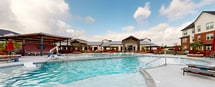 This expansive outdoor pool area is designed as a resort-style amenity space, offering a comprehensive range of recreational and relaxation features. At its center sits a large, curved swimming pool with crystal-clear turquoise water, complemented by a separate hot tub and gentle water features along the pool's edge. The surrounding concrete sundeck is generously appointed with lounge chairs and red-accented umbrellas, providing ample space for sunbathing and outdoor leisure. A standout architectural feature is the covered pavilion, constructed with steel posts and a warm wood-paneled ceiling equipped with ceiling fans, offering shaded dining and gathering space with tables and chairs. A wall-mounted television enhances the entertainment options beneath the pavilion. Additional shaded pergola structures with warm wood siding are positioned throughout the deck, creating intimate seating nooks. Raised brick planters with tropical palms and ornamental grasses add greenery and visual interest throughout the space. The perimeter of the pool area is framed by multi-story residential buildings featuring a combination of red brick and white horizontal siding, with large glass windows and doors opening onto covered exterior walkways furnished with seating. The overall ambiance is bright, modern, and community-focused.