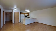 This single-floor unit is designed with an open-concept layout that seamlessly connects the kitchen and living area, creating a practical and cohesive living environment. The kitchen is equipped with dark wood cabinetry providing ample storage, a white refrigerator, an electric range, and an integrated sink set within a peninsula-style counter. The peninsula serves as a natural divider between the kitchen and the adjacent living space, offering additional workspace and casual seating potential.

Light hardwood flooring extends throughout the main area, lending warmth and visual continuity to the space. The neutral, cream-toned walls enhance the sense of openness and pair well with the wood accents found in both the cabinetry and flooring. Overhead lighting in the kitchen includes a fluorescent fixture that ensures ample task lighting, while a flush-mount ceiling light serves the broader living area.

A paneled interior door is visible near the kitchen, likely providing access to additional areas of the unit. The overall layout is straightforward and efficient, making good use of available space while maintaining a clean, functional aesthetic suited for everyday living.