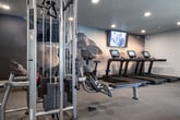 This fitness center in Boise, Idaho, offers a thoughtfully designed, single-floor layout that accommodates a wide range of workout styles and fitness goals. The main exercise room is generously proportioned, featuring textured ceilings with recessed lighting that keeps the space bright and inviting. Large windows along one wall flood the area with natural light and provide outdoor views, creating an open, airy atmosphere. The main floor is equipped with multiple cardio machines, including treadmills and stationary bikes, positioned in front of wall-mounted televisions and a striking mountain-themed mural that adds a dynamic visual element. A dedicated strength training section features cable machines, a multi-rack system, adjustable benches, and a tiered dumbbell rack. Full-length mirrors and a wall-mounted ballet barre line one side of the room, reflecting the space and supporting stretching and flexibility training. A sliding barn-style door leads into a separate functional training room with light wood flooring, offering an organized selection of kettlebells, medicine balls, battle ropes, resistance bands, a BOSU balance trainer, and a full dumbbell rack. Exposed ductwork adds an industrial aesthetic to this secondary space. Together, the two areas provide a comprehensive and versatile environment suited for cardio, strength, and functional fitness training.