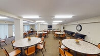 This single-floor communal space is designed to accommodate group dining, social gatherings, and multipurpose events. The main dining area is expansive and open, furnished with numerous round tables topped with granite-style surfaces and surrounded by padded orange chairs, providing comfortable seating for a large number of occupants. Flush fluorescent lighting panels illuminate the entire space evenly, contributing to a bright and welcoming atmosphere. Large windows and sliding glass doors along one wall allow natural light to filter in while offering views of the outdoors. A wall-mounted flat-screen television serves as a focal point at one end of the room, making the space suitable for presentations or group viewing. An adjoining kitchenette is well-appointed with warm wood cabinetry, granite-style countertops, hardwood flooring, and a full suite of stainless steel appliances including a refrigerator, dishwasher, wall ovens, and a microwave. A secondary area near the rear of the space provides additional seating alongside built-in bench seating and houses utility carts, suggesting functional storage and event support. Wall-mounted mini-split HVAC units ensure climate control throughout. The space is designed with practicality and community use in mind, offering a clean, organized, and accommodating environment.