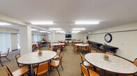 This single-floor communal space is designed to accommodate group dining, social gatherings, and multipurpose events. The main dining area is expansive and open, furnished with numerous round tables topped with granite-style surfaces and surrounded by padded orange chairs, providing comfortable seating for a large number of occupants. Flush fluorescent lighting panels illuminate the entire space evenly, contributing to a bright and welcoming atmosphere. Large windows and sliding glass doors along one wall allow natural light to filter in while offering views of the outdoors. A wall-mounted flat-screen television serves as a focal point at one end of the room, making the space suitable for presentations or group viewing. An adjoining kitchenette is well-appointed with warm wood cabinetry, granite-style countertops, hardwood flooring, and a full suite of stainless steel appliances including a refrigerator, dishwasher, wall ovens, and a microwave. A secondary area near the rear of the space provides additional seating alongside built-in bench seating and houses utility carts, suggesting functional storage and event support. Wall-mounted mini-split HVAC units ensure climate control throughout. The space is designed with practicality and community use in mind, offering a clean, organized, and accommodating environment.
