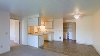 This single-floor unit offers an open, functional layout where the kitchen flows seamlessly into the living area. The kitchen is equipped with white cabinetry, a white electric range, a refrigerator, and a double-basin sink positioned along the counter. A peninsula extends from the kitchen, providing additional workspace and serving as a natural divider between the kitchen and living space. The flooring in the kitchen transitions from tile to carpet in the living area, clearly defining each zone. Overhead, a ceiling fan with a light fixture serves the adjoining living space, while under-cabinet lighting illuminates the kitchen work surfaces. Toward the back of the unit, an open utility closet houses a washer and dryer, offering convenient in-unit laundry access. Built-in shelving within the utility area provides additional storage. The walls are finished in a neutral white palette throughout, and the overall layout is straightforward and practical. A wall-mounted heater is visible near the base of the wall, ensuring climate control within the space. The unit presents a clean, no-frills design suited for everyday living.