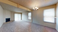 Located at 9110 Broadway St in San Antonio, Texas, this single-floor residential unit offers a practical and functional layout designed for comfortable everyday living. The open living and dining area is a central feature, finished with neutral-toned carpet underfoot and a textured ceiling overhead. A classic chandelier anchors the dining space, while a pass-through window connects it to the kitchen, creating a sense of openness. Vertical blinds cover a sliding glass door that brings in natural light and provides outdoor access, complemented by additional windows fitted with horizontal blinds throughout. The kitchen is efficiently arranged in a U-shape, featuring white cabinetry, dark countertops, a double-basin sink, a coil-top range, and a dishwasher. Tile flooring runs through the kitchen and bathroom areas. The bedroom is a well-proportioned, carpeted room with a ceiling fan and ample wall space. The bathroom includes built-in cabinetry for storage, tile flooring, and a private water closet. A carpeted stairway with a decorative wall-mounted lantern provides access between levels. The unit maintains a consistent, neutral color palette throughout, lending a clean and cohesive ambiance.