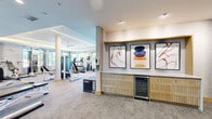 Located at 2275 Chester St in Aurora, CO, this single-floor fitness center offers a comprehensive and thoughtfully designed wellness facility. The space is divided into two primary zones that together deliver a full-spectrum exercise experience.

The main gym area features a bright, open layout with a tray ceiling accented by recessed and indirect lighting, along with ceiling fans for comfort. Large windows line the perimeter, flooding the space with natural light and offering outdoor views. This zone houses a wide array of cardio equipment — including treadmills and ellipticals — as well as a full complement of strength machines, free weights, a barbell rack, and benches. Built-in shelving provides additional storage and organization.

A separate cycling studio offers a distinct atmosphere, distinguished by rich hardwood flooring, a striking coffered ceiling with warm wood trim, and floor-to-ceiling glass garage doors that open to the outdoors. Wall-mounted mirrors with arched frames amplify the sense of space, and a projector system supports instructor-led classes. Kettlebells, medicine balls, and resistance equipment round out the room's functionality.

A transitional lounge area connects the zones, featuring a sleek built-in credenza with a wine cooler, white countertop, and a trio of framed abstract artworks — adding a refined, boutique aesthetic to the overall facility.