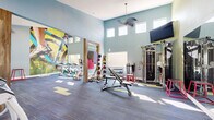 Located at 3223 W Ina Rd in Tucson, Arizona, this fitness center offers a thoughtfully designed workout facility across a single open floor. The space is defined by its soaring ceilings, which lend an airy, expansive feel, and a row of clerestory windows that usher in abundant natural light throughout the day. Ceiling fans help maintain comfortable airflow during workouts.

The interior is painted in a calming combination of soft blue and sage green, creating an energizing yet approachable atmosphere. A large, vivid cycling-themed wall mural serves as a bold focal point, reinforcing the active spirit of the space. Durable, blue-toned carpet tiles with yellow accent patterns cover the open floor, providing cushioning and visual interest.

The equipment layout is well-organized and comprehensive. A dedicated cardio zone houses multiple treadmills and elliptical machines, while a separate area accommodates spin bikes. Strength training is supported by a multi-function cable machine, an adjustable weight bench, a full dumbbell rack, and a medicine ball rack. Large wall mirrors expand the sense of space and assist with form. A wall-mounted flat-screen TV enhances the workout experience. French doors on one end provide direct outdoor access, connecting the fitness center to the surrounding community.