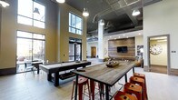 This single-floor communal lounge showcases an industrial-modern aesthetic defined by soaring double-height ceilings with exposed ductwork and pendant lighting. Large floor-to-ceiling windows along one wall flood the space with natural light while offering views of the surrounding exterior. The open-plan layout is divided into distinct activity zones that flow seamlessly into one another. A full-size billiards table anchors the games area, accompanied by a substantial reclaimed wood high-top communal table surrounded by red metal bar stools, adding a rustic contrast to the otherwise industrial framework. Structural columns with a clean white finish help delineate the space without interrupting sightlines. Toward the rear, a dedicated lounge area features a wall-mounted flat-screen television set against a striking reclaimed wood accent wall, creating a warm, textured focal point. The light-toned flooring throughout contributes to the bright, airy atmosphere. The combination of raw materials, intentional lighting, and varied seating options makes this a versatile, well-appointed social space suited for both casual gatherings and recreational activities.
