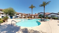 This inviting outdoor amenity area serves as the communal centerpiece of a multi-building residential community, designed with a Southwestern architectural aesthetic. The buildings feature stucco exteriors, red tile rooflines, and teal accent doors, creating a cohesive and warm visual character throughout the property.

At the heart of the space is a generously sized freeform swimming pool with crystal-clear blue water, complemented by an adjacent spa. A covered pavilion with columns provides a shaded gathering area, while the surrounding concrete pool deck is furnished with blue lounge chairs and dining seating, ideal for outdoor relaxation. Tall palm trees and mature shade trees are thoughtfully positioned throughout the space, offering natural relief from the sun.

The landscaping follows a desert-friendly design, incorporating gravel ground cover, ornamental shrubs, yucca plants, and drought-tolerant greenery that add texture and color without requiring intensive upkeep. The pool area is enclosed by a sleek dark metal fence, providing both security and an open sightline to the surrounding buildings and grounds.

Stairways and balconies are visible on the multi-story residential buildings, and the overall layout emphasizes connectivity between the outdoor amenity space and the living quarters. The property enjoys abundant natural sunlight under wide-open skies, creating a bright and welcoming atmosphere throughout.