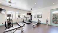 Located in Escondido, California, this fitness center is thoughtfully designed across two exercise rooms, offering a comprehensive workout environment. The space features a clean, modern aesthetic with crisp white walls accented by teal/aqua trim and door frames, durable rubberized flooring, and recessed lighting complemented by ceiling fans for optimal air circulation.

The main exercise room is generously proportioned and outfitted with a variety of cardio and strength equipment, including treadmills, a stationary bike, rowing machines, and a dumbbell rack with an adjustable weight bench. Large floor-to-ceiling mirrors line the walls, visually expanding the space and allowing users to monitor their form. A wall-mounted television provides entertainment during workouts. Multiple windows with blinds allow abundant natural light to fill the room while maintaining privacy, and a set of teal-framed glass double doors connects the space to the outdoors.

The second, more intimate exercise room serves as a dedicated stretching and functional training area, equipped with stability balls on a storage rack and a padded workout bench. Natural light filters in through windows, creating a calm environment ideal for flexibility training or low-impact exercise. Together, the two rooms provide a well-rounded, amenity-rich fitness facility suited for a variety of workout routines.