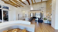 Located at 4320 Commons Dr W in Destin, Florida, this single-floor gathering space blends coastal charm with modern design in a seamlessly open layout. The interior is immediately defined by dramatic vaulted ceilings adorned with exposed rustic wood beams, creating a warm yet contemporary atmosphere that draws the eye upward. Warm hardwood-style flooring runs throughout, grounding the bright, airy aesthetic established by a predominantly white color palette across the walls, cabinetry, and furnishings. A full kitchen and bar area anchors one side of the space, complete with white shaker-style cabinetry, a large counter with bar stool seating, and pendant lighting that cascades elegantly from above. The lounge area features a generous curved white sofa and accent chairs arranged to foster social interaction, with organic wood side tables adding a natural touch. A sculptural statement light fixture serves as a focal point near the entrance, complementing the layered pendant lighting throughout. Large glass doors and windows at the far end invite abundant natural light and provide views to the exterior, enhancing the open, welcoming character of the space. The overall ambiance is refined yet relaxed — a well-appointed communal environment ideal for gatherings and everyday social connection.