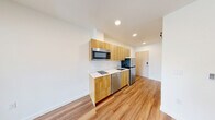 Located in Seattle, WA, this efficiently designed one-bedroom, one-bathroom unit offers a thoughtful layout that maximizes every square foot. The open-concept kitchen and living area form the heart of the space, featuring warm wood-toned cabinetry, white quartz-style countertops, and a full suite of stainless steel appliances including a refrigerator, dishwasher, over-the-range microwave, and a cooktop. Recessed lighting throughout keeps the space bright and modern, while light-toned hardwood-style flooring flows seamlessly from room to room, adding warmth and visual continuity. A sliding barn door with sleek hardware serves as a stylish and space-saving divider, enhancing the modern aesthetic. The living area is clean and open, with crisp white walls and a frosted-glass interior door that allows natural light to filter through from the bedroom. The bedroom is a cozy, well-defined retreat with access to a dedicated closet. The bathroom continues the clean design language, featuring matching wood cabinetry, a white vanity countertop, and wood-look flooring. The overall ambiance is contemporary and minimalist, making smart use of a compact footprint for efficient urban living in Seattle.