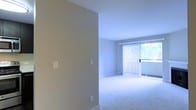 This single-floor space offers a clean, minimalist layout designed for comfortable everyday living. The interior is finished with smooth, neutral-toned walls that create a calm and cohesive atmosphere throughout. Plush wall-to-wall carpeting adds warmth underfoot, contributing to a quiet and comfortable feel. The living area is a standout feature, anchored by a large sliding glass door dressed with vertical blinds that allow for flexible light control. This expansive glazed opening invites generous natural light into the space and provides direct access to an outdoor area, framing views of surrounding greenery. The transition between rooms is defined by a thoughtfully placed partial wall, subtly dividing the entry corridor from the main living area while maintaining an open and airy sense of flow. Light switch panels are neatly integrated into the walls, and the white baseboard trim provides a crisp finish against the carpeted floors. Overall, the space presents a straightforward, functional design with an emphasis on natural light, neutral finishes, and an open, uncluttered layout that offers a versatile and welcoming environment.