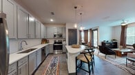 Located at 2300 Laurel Rd E in Venice, Florida, this single-story residence offers a well-appointed layout designed for comfortable living. The open-concept living area features wood-look flooring, a ceiling fan, and generous natural light streaming through large windows dressed in warm earth-toned curtains. A curated gallery of woven basket wall art lends a coastal, bohemian character to the space, complemented by a tufted sofa, accent chairs, and a solid wood coffee table.

The kitchen is a standout feature, showcasing grey shaker-style cabinetry, stainless steel appliances, white quartz countertops, a subway tile backsplash, and a curved peninsula with bar seating. Pendant lighting adds a refined touch overhead. Adjacent to the kitchen, a dedicated dining nook provides an intimate setting with a round reclaimed wood table surrounded by woven rattan chairs and natural light from nearby windows.

The bedroom is a serene retreat with carpet flooring, large windows overlooking lush greenery, and tasteful neutral décor. The bathroom is finished with large-format grey tile in a glass-enclosed shower, wood-look flooring, and a clean white vanity with quartz countertops. Together, these spaces create a cohesive, stylish home with a relaxed Florida aesthetic.