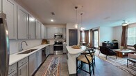 Located at 2300 Laurel Rd E in Venice, Florida, this single-story residence offers a well-appointed layout designed for comfortable living. The open-concept living area features wood-look flooring, a ceiling fan, and generous natural light streaming through large windows dressed in warm earth-toned curtains. A curated gallery of woven basket wall art lends a coastal, bohemian character to the space, complemented by a tufted sofa, accent chairs, and a solid wood coffee table.

The kitchen is a standout feature, showcasing grey shaker-style cabinetry, stainless steel appliances, white quartz countertops, a subway tile backsplash, and a curved peninsula with bar seating. Pendant lighting adds a refined touch overhead. Adjacent to the kitchen, a dedicated dining nook provides an intimate setting with a round reclaimed wood table surrounded by woven rattan chairs and natural light from nearby windows.

The bedroom is a serene retreat with carpet flooring, large windows overlooking lush greenery, and tasteful neutral décor. The bathroom is finished with large-format grey tile in a glass-enclosed shower, wood-look flooring, and a clean white vanity with quartz countertops. Together, these spaces create a cohesive, stylish home with a relaxed Florida aesthetic.