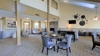 This two-story space impresses with its dramatically tall ceilings and open, airy layout. A central support column anchors the room while emphasizing the generous vertical height, with upper-level windows allowing natural light to filter down into the interior. The main level is thoughtfully arranged into distinct functional zones. A dining area near the center features a round dark-topped table surrounded by upholstered gray chairs, offering a comfortable spot for casual gatherings. Adjacent to this, a lounge area is furnished with a neutral-toned sofa, accent chairs, and decorative pillows, creating a relaxed seating arrangement. A fireplace serves as a focal point in the background, flanked by additional seating and framed by a refined surround. A wall-mounted flat-screen television is positioned nearby for added convenience. The flooring throughout features a neutral-toned carpet that complements the warm beige and gray color palette of the space. Decorative accents, including sculptural pieces and a table lamp, add refined touches throughout. French doors at the far end provide access to the exterior, introducing natural views and enhancing the overall sense of openness.