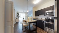 This single-floor residence offers a thoughtfully designed open-concept layout that seamlessly connects the kitchen, dining, and living areas. The kitchen is a standout feature, showcasing rich dark cabinetry that contrasts beautifully with light granite countertops. A central kitchen island with bar seating provides a casual dining option and additional prep space, while pendant lighting above adds a modern, polished touch. Stainless steel appliances and a double-basin sink with granite surround complete the kitchen's functional and refined aesthetic. Wood-style flooring flows consistently throughout the space, lending warmth and visual continuity. The open living and dining area beyond the kitchen is bright and inviting, benefiting from abundant natural light streaming through large windows and a sliding glass door, which also provides access to an outdoor area. A ceiling fan in the living space adds both comfort and a finishing detail. Neutral wall tones keep the palette clean and versatile, while the overall layout promotes a comfortable and connected living experience. The combination of modern finishes, natural light, and cohesive design makes this a well-appointed and functional single-floor living space.