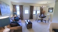 This single-floor space features an open-concept layout that connects the living and dining areas in a cohesive, functional design. The living area is anchored by a plush sofa adorned with teal accent pillows and centered around a distinctive ornate coffee chest, adding a decorative focal point to the room. Neutral-toned carpeting runs throughout, contributing to a warm and inviting atmosphere.

Large windows on multiple walls flood the space with natural light, offering views of the surrounding outdoor scenery. Rich, dark floral curtains with roller shades provide both style and light control, creating a layered window treatment that adds depth and elegance to the room.

The dining area transitions seamlessly from the living space and is furnished with a dark wood round table surrounded by upholstered white chairs, complemented by a refined chandelier overhead that introduces a touch of sophistication. A decorative floral arrangement serves as a centerpiece on the dining table. A white entry door is centrally positioned along the front wall, enhancing the open and airy feel of the overall space.
