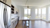 This two-bedroom, two-bathroom residence showcases a thoughtfully designed open-concept layout that seamlessly connects the kitchen and living areas. The kitchen is outfitted with sleek, light-toned cabinetry featuring open upper shelving, a undermount sink set into white quartz countertops, and stainless steel appliances including a built-in microwave and range. A central island provides additional prep space and casual seating potential, accented by stylish pendant lighting suspended from the ceiling above.

The space benefits from an abundance of natural light, courtesy of large floor-to-ceiling windows that frame sweeping views of a lush, tree-lined cityscape. The warm-toned hardwood flooring runs throughout, adding richness and continuity to the clean, contemporary aesthetic. White walls and high ceilings amplify the sense of openness, while recessed and track lighting provide flexible illumination options. The overall ambiance is bright, airy, and modern, with a neutral palette that offers a versatile backdrop for a variety of interior styles.
