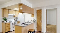 This single-floor one-bedroom unit offers a thoughtfully laid-out living space designed for both comfort and functionality. The open-concept living area features light wood-style flooring throughout, crisp white walls, and a sliding glass door that floods the room with natural light while providing outdoor access. The kitchen is a standout feature, designed in a galley style with warm wood-toned cabinetry lining both walls, gray laminate countertops, and white appliances including a range and refrigerator. A skylight panel overhead brightens the kitchen interior, while a peninsula with a built-in sink and bar seating connects seamlessly to the living area, making it ideal for casual dining or entertaining. Adjacent to the kitchen, a solid wood door leads to the bedroom and closet. The spacious bedroom is finished with plush carpet flooring and features a window that allows in natural light. A dedicated walk-in closet off the bedroom provides generous storage, complete with wire shelving. The unit's clean, neutral palette and practical layout create an inviting and functional living environment suited for everyday living.