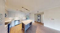 Located at 6460 Main Street in Miami Lakes, FL, this well-appointed 1-bedroom, 2-bathroom residence offers a thoughtfully designed layout across a single floor. The open-concept kitchen and living area serves as the heart of the home, featuring light gray flat-panel cabinetry, a mosaic tile backsplash, stainless steel appliances including a gas range, built-in microwave, refrigerator with ice dispenser, and dishwasher. A freestanding center island with a wood-toned base and white countertop provides additional prep and dining space. Track lighting illuminates the kitchen and living area, which flows over warm-toned hardwood-style flooring. A stacked washer and dryer unit is conveniently integrated into the main living space. The spacious primary bedroom features plush carpet, a ceiling fan, and direct access to a walk-in closet with built-in shelving and dual hanging rods. Both bathrooms are finished with elongated white vanities, modern fixtures, large mirrors, and backlit vanity lighting, with natural light visible in the primary bath. The clean, neutral color palette and contemporary finishes throughout create a bright, modern ambiance well-suited to South Florida living.