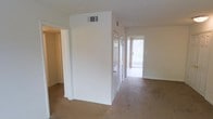This single-floor space is designed with a straightforward, functional layout that connects multiple rooms through an open corridor. The interior is finished with wall-to-wall carpeting in a warm beige tone, contributing to a comfortable and cohesive aesthetic throughout. The walls are painted in a clean, neutral white, creating a bright and airy atmosphere that complements the natural light filtering in from what appears to be a sliding glass door or large window at the end of the hallway, fitted with vertical blinds that allow for adjustable light control.

The corridor serves as a central passage, with built-in closet doors visible along the wall, providing practical storage solutions within the living space. HVAC vents are positioned in the ceiling, ensuring climate control throughout the unit. The doorways leading to adjacent rooms are generously sized, supporting easy movement between spaces.

The overall ambiance is clean and understated, with an emphasis on light tones and simplicity. The glimpse of an outdoor area through the far window suggests access to a patio or balcony, adding an element of openness to the interior. The space is move-in ready and well-suited for straightforward, comfortable living.