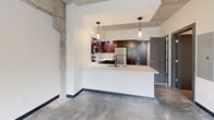 Located at 112 Wayne Ave in Dayton, Ohio, this single-floor unit showcases an industrial-modern aesthetic that blends raw architectural elements with contemporary finishes. The space is defined by polished concrete floors that run throughout, lending a cohesive, loft-inspired character to every room.

The kitchen serves as a central focal point, featuring rich dark-wood cabinetry extending to the ceiling for ample storage, white countertops, and a full suite of stainless steel appliances including a refrigerator, range, and built-in microwave. A peninsula with an integrated sink provides additional workspace and a natural divide between the kitchen and the main living area. Pendant lighting hangs above, adding warmth and definition to the space.

Exposed concrete columns punctuate the interior, reinforcing the industrial character of the building. The main living area benefits from a large window that introduces natural light and outdoor views, complemented by an overhead pendant fixture and exposed concrete ceiling structure above. A hallway connects the areas, where raw concrete wall surfaces contrast with clean white walls. Dark-framed interior doors with brushed metal hardware add refined detail throughout.