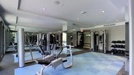 This fitness center is a thoughtfully designed workout facility that caters to a wide range of exercise needs. The space is outfitted with a robust selection of strength-training machines, including cable systems and multi-function weight stations, providing users with everything needed for a full-body workout. The layout is open and organized, allowing for easy movement between equipment. Floor-to-ceiling mirrors line the walls, creating a sense of expansiveness while allowing users to monitor their form during exercise. The flooring features durable, cushioned tiles in a blue and grey pattern, offering both comfort underfoot and impact absorption — a practical choice for a high-use fitness environment. Recessed lighting in the white ceiling ensures the space is bright and well-lit throughout. The overall ambiance is clean, professional, and functional, reflecting a facility maintained to a high standard. Large windows allow natural light to filter into the space, further enhancing the energizing atmosphere. Whether for strength training or general conditioning, this fitness center provides a complete and comfortable environment for users of all fitness levels.