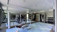 This fitness center is a thoughtfully designed workout facility that caters to a wide range of exercise needs. The space is outfitted with a robust selection of strength-training machines, including cable systems and multi-function weight stations, providing users with everything needed for a full-body workout. The layout is open and organized, allowing for easy movement between equipment. Floor-to-ceiling mirrors line the walls, creating a sense of expansiveness while allowing users to monitor their form during exercise. The flooring features durable, cushioned tiles in a blue and grey pattern, offering both comfort underfoot and impact absorption — a practical choice for a high-use fitness environment. Recessed lighting in the white ceiling ensures the space is bright and well-lit throughout. The overall ambiance is clean, professional, and functional, reflecting a facility maintained to a high standard. Large windows allow natural light to filter into the space, further enhancing the energizing atmosphere. Whether for strength training or general conditioning, this fitness center provides a complete and comfortable environment for users of all fitness levels.