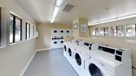 This shared laundry facility is designed with functionality and practicality in mind. The space is laid out as a single elongated room with neutral-toned walls in soft beige and gray, complemented by clean white trim and gray carpet flooring throughout. Overhead fluorescent lighting ensures the entire area is consistently well-lit.

The laundry area is well-equipped with a row of front-loading washing machines along one side, paired with stacked dryer units along the back wall, providing ample capacity for multiple users simultaneously. Posted laundry facility rules are displayed on the wall for resident reference.

A built-in folding counter with a dark speckled laminate surface runs along one side of the room, offering generous workspace for sorting and folding laundry. Below the counter, open cubbies provide additional organizational storage. A utility sink with a dark countertop is also integrated into the workspace, adding further convenience.

Several windows are positioned along the walls, allowing natural light to filter into the space and creating a more open, airy atmosphere. A private door provides a dedicated entry and exit point. Overall, the facility is clean, spacious, and thoughtfully arranged to accommodate the laundry needs of multiple users efficiently.