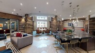Located at 3202 W Colfax Ave in Denver, Colorado, this single-floor communal amenity space offers a sophisticated and well-appointed environment designed for both socializing and relaxation. The interior design blends contemporary elegance with rustic warmth, prominently featuring reclaimed wood-clad columns and accent walls throughout.

The space is organized into several distinct zones that flow seamlessly into one another. A central lounge area anchors the layout, furnished with plush seating and a striking marble-surround fireplace flanked by built-in navy bookshelves and a wall-mounted television. Adjacent to this is a sleek bar and entertainment area, complete with a dramatic dark marble countertop, gold-accented bar stools, under-counter refrigeration, and multiple mounted flat-screen televisions set against a dark tile backsplash.

Pendant lighting clusters add a warm, layered glow throughout, complementing the recessed ceiling lights above polished tile flooring laid in a herringbone pattern. A separate lounge zone features floor-to-ceiling windows that flood the space with natural light and offer views of the surrounding courtyard. This area includes a long communal table with upholstered seating, set atop herringbone-patterned carpet in cool blue tones, ideal for collaborative or casual use.
