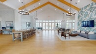 Located at 3600 Courtney Dr in Panama City, Florida, this single-story clubhouse facility offers an expansive, open-concept layout designed for both community gathering and everyday functionality. The architectural centerpiece is a dramatic vaulted ceiling accented by natural wood beams, which lend warmth and character to the bright, airy interior. Large-scale ring chandeliers hang throughout, complementing the recessed lighting and contributing to the space's polished yet welcoming ambiance.

Light hardwood flooring flows continuously across the main gathering areas, reinforcing the sense of openness and cohesion. A wall of French doors and transom windows along the rear of the primary hall floods the space with natural light and provides direct views and access to the outdoor pool area. Multiple seating arrangements accommodate a range of uses, from casual lounging to social gatherings, while a game area featuring a pool table adds a recreational element.

A dedicated kitchenette area showcases sea-glass tile backsplash, white quartz countertops, hexagonal tile flooring, and a built-in beverage station. Private office space is also present, featuring tall ceilings, sheer-draped windows with outdoor views, and refined pendant lighting. Throughout, a coastal-inspired palette of soft whites, blues, and natural textures reflects the Florida Gulf Coast setting.