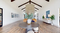 This expansive single-floor communal space offers an impressive open-concept layout encompassing a living room, dining area, and kitchen. The architectural centerpiece is a dramatic vaulted ceiling accented by bold, dark-stained exposed wood beams that run the length of the space, creating a striking visual contrast against the crisp white walls and adding a sense of grandeur. A statement fireplace wall with paneled tile detail anchors the living area, which is outfitted with plush blue seating, accent chairs, and a modern circular chandelier overhead. Wood-look tile flooring flows seamlessly throughout, unifying each zone. The kitchen features bold navy-blue cabinetry with brass hardware, a decorative tile backsplash, open shelving, a built-in beverage refrigerator, and a dedicated coffee station — designed for both functionality and style. The dining area includes a curved banquette with marble-topped bistro tables, framed by floor-to-ceiling windows dressed in warm terracotta drapes that invite an abundance of natural light and outdoor views. A glass-enclosed leasing office with black-framed doors adds a modern, professional element. Curated artwork, lush indoor plants, and thoughtful furnishings complete the welcoming, contemporary ambiance of this well-appointed space.