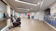 Located at 222 W Boston St in Chandler, AZ, this contemporary fitness center offers a well-appointed, single-floor space designed to support a variety of workout routines. The open layout provides ample room for both cardio and functional training, with light wood-toned flooring throughout that gives the space a clean, modern aesthetic.

A row of smart stationary spin bikes lines one side of the room, each equipped with individual display screens and positioned in front of large windows that allow natural light to filter in and offer views of the surrounding exterior. The open floor area beyond the bikes provides generous space for stretching, floor exercises, or group fitness activities.

One of the most striking design features is a large, abstract wall mural rendered in rich blues and grays, which anchors the space with an artistic focal point and adds personality to the otherwise crisp, neutral interior. A wall-mounted flat-screen display is integrated within the mural, ideal for guided workouts or media.

Recessed and linear LED ceiling lights provide consistent, even illumination throughout. A dedicated storage rack holds stability balls and resistance accessories, while a full-length mirror along one wall supports form-checking during exercise. A conveniently located restroom and dual water fountain station complete the amenities, making this a fully self-contained fitness facility.