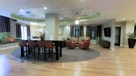 This single-floor community space offers a thoughtfully designed open-plan layout that seamlessly blends comfort and functionality. The interior is anchored by rich hardwood flooring that flows throughout, providing a warm and inviting foundation for the entire area.

The space is divided into two distinct seating zones. A high-top dining area features a dark wood bar-height table surrounded by matching upholstered chairs, accented by a decorative floral arrangement that adds a refined touch. Adjacent to this is a relaxed lounge area centered on a circular area rug, where a cluster of terra cotta-toned armchairs invites casual conversation and relaxation.

Architectural interest is introduced through varied ceiling heights, with recessed lighting and a stylish pendant fixture providing layered illumination. Subtle green accent lighting along the ceiling adds a contemporary flair and visual depth to the room. The walls are painted in soft yellow-green tones, complemented by floor-to-ceiling curtain panels in rich brown, which frame the large windows and allow for natural light. A large flat-screen television is mounted on one wall, adding a multimedia element to the lounge. The overall ambiance is modern yet comfortable, making this a versatile space well-suited for both social gatherings and quiet relaxation.