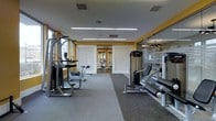 This modern fitness center is designed to support a comprehensive workout experience within a single-level layout. The space is thoughtfully organized to accommodate a variety of exercise equipment, including strength-training machines and cardio apparatuses, arranged to allow for comfortable movement and use throughout the room.

The interior features durable rubber flooring suited for high-traffic athletic use, while bright yellow accent walls add energy and visual interest to the environment. The ceiling is finished in white and outfitted with linear fluorescent lighting strips that provide ample, even illumination throughout the space.

A standout design element is the glass partition wall along one side of the room, which visually opens the space and connects it to an adjacent area, enhancing the overall sense of spaciousness. Decorative perforated panels are incorporated into the design, adding texture and a modern aesthetic.

Natural light filters in through windows along the perimeter, complementing the artificial lighting and contributing to a bright, welcoming atmosphere. The layout is open and uncluttered, ensuring ease of navigation between equipment stations. Overall, this fitness center offers a clean, functional, and well-designed environment suited for a range of fitness activities.