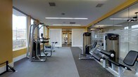 This modern fitness center is designed to support a comprehensive workout experience within a single-level layout. The space is thoughtfully organized to accommodate a variety of exercise equipment, including strength-training machines and cardio apparatuses, arranged to allow for comfortable movement and use throughout the room.

The interior features durable rubber flooring suited for high-traffic athletic use, while bright yellow accent walls add energy and visual interest to the environment. The ceiling is finished in white and outfitted with linear fluorescent lighting strips that provide ample, even illumination throughout the space.

A standout design element is the glass partition wall along one side of the room, which visually opens the space and connects it to an adjacent area, enhancing the overall sense of spaciousness. Decorative perforated panels are incorporated into the design, adding texture and a modern aesthetic.

Natural light filters in through windows along the perimeter, complementing the artificial lighting and contributing to a bright, welcoming atmosphere. The layout is open and uncluttered, ensuring ease of navigation between equipment stations. Overall, this fitness center offers a clean, functional, and well-designed environment suited for a range of fitness activities.