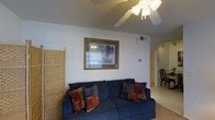 This single-floor space features a welcoming living area as its centerpiece. The room is anchored by a plush navy blue sofa adorned with colorful patterned throw pillows, offering a comfortable and inviting seating arrangement. A ceiling fan with a light fixture overhead provides both ventilation and ambient lighting, complementing the textured ceiling. The neutral-toned walls are accented by a gold-framed artwork, adding a decorative focal point to the space. A woven rattan room divider on one side introduces a natural texture, subtly defining the area. Light-colored carpeting runs throughout, contributing to a warm and casual atmosphere. A wall-mounted HVAC vent ensures climate control throughout the space. A hallway visible in the background leads to additional areas of the home, where track lighting and a glimpse of a wooden chair hint at further living spaces beyond. The overall ambiance is relaxed and functional, with a straightforward, comfortable layout suited for everyday living.
