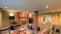 This single-floor space is designed for comfort and casual entertaining, offering a cohesive and welcoming atmosphere throughout. The main living area serves as the focal point, anchored by a tile-surround fireplace set against a striking terra cotta accent wall that adds warmth and character to the room. A structural column finished in a coordinating tone provides an architectural detail that subtly defines the space without disrupting its open feel. Recessed lighting and track lighting illuminate the area evenly, complementing the neutral carpet flooring. The living area is furnished with rich, dark leather sofas and matching ottomans arranged around a glass-top coffee table, with a decorative area rug grounding the seating arrangement. Wall-mounted artwork and decorative plates add personality and a curated aesthetic to the space. A dark wood media cabinet provides additional storage and display options. Toward the rear, a set of frosted glass double doors leads to an additional room, offering flexible use of the space. The overall ambiance is warm, grounded, and thoughtfully appointed, making it a comfortable and functional retreat.