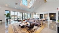 Located in San Diego, CA, this single-story community clubhouse at 7231 Colchester Ct delivers a striking blend of contemporary and coastal design. The open-concept interior is anchored by soaring vaulted ceilings clad in white shiplap, allowing natural light to pour in through oversized skylights and floor-to-ceiling black-framed windows. Polished concrete floors stretch throughout, reflecting light and lending a clean, modern aesthetic.

The main lounge area offers generous seating with plush sectional sofas, leather accent chairs, and layered vintage-style rugs, creating an inviting social hub. A dramatic feature wall clad in dark wood paneling houses a linear fireplace and a wall-mounted television, flanked by built-in shelving displaying curated décor. Adjacent to this, a long communal dining table surrounded by woven leather counter stools provides an ideal setting for gathering, with direct views of the palm-lined pool courtyard beyond the glass walls.

A dedicated workspace or leasing area is enclosed by glass-panel partitions, maintaining visual openness while offering a defined zone for business. A kitchenette with open shelving and bar seating adds further functionality. Throughout, the space is finished with a sculptural chandelier, thoughtful artwork, and lush indoor plants, reinforcing the relaxed yet refined Southern California atmosphere.