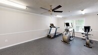 Located at 1030 Atlas Rd in Columbia, SC, this fitness center at Salon Place offers a clean, well-appointed exercise space designed to meet the essential workout needs of its users. The room features durable speckled rubber flooring throughout, providing a safe and resilient surface ideal for exercise equipment and physical activity.

The space is equipped with a variety of cardio and strength-training machines, including a treadmill, an elliptical trainer, a recumbent stationary bike, and a multi-station cable strength machine with a weight rack. A wall-mounted flat-screen television is positioned for easy viewing during workouts, enhancing the overall exercise experience.

Bright fluorescent lighting and a ceiling fan with a light fixture ensure the space remains well-lit and comfortably ventilated. Crown molding and a chair-rail style wainscot trim along the walls lend a polished, finished look to the interior. A textured ceiling adds a classic touch throughout. Natural light filters in through a window, offering a pleasant view of the surrounding greenery.

Fitness center rules are posted near the entrance, and a fire safety alarm is visibly installed on the wall, reflecting attention to safety standards. The space maintains a clean, neutral color palette of warm beige tones, creating an inviting and focused environment for exercise.