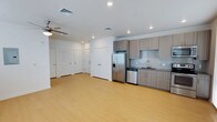 This single-floor space offers a clean, modern design with an open-concept layout that seamlessly connects the living area and kitchen. The light hardwood flooring extends throughout, brightening the interior and contributing to a spacious, airy feel. Recessed lighting is installed across the white ceiling, complemented by a ceiling fan that adds both function and a casual touch to the living area.

The kitchen is outfitted with contemporary flat-front cabinetry in a warm taupe wood finish, paired with a subtle tile backsplash in a neutral grey tone. Stainless steel appliances include a French door refrigerator with a bottom freezer and ice dispenser, a dishwasher, and an undermount sink with a modern faucet. Upper and lower cabinetry provide ample storage while maintaining a sleek, uncluttered aesthetic.

Along the opposite wall, multiple white panel doors lead to additional rooms or closet spaces, keeping the layout organized and functional. The overall ambiance is contemporary and minimalist, with a neutral color palette of whites and warm wood tones that creates a welcoming, versatile environment suitable for a variety of furnishing styles.