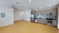 This single-floor space offers a clean, modern design with an open-concept layout that seamlessly connects the living area and kitchen. The light hardwood flooring extends throughout, brightening the interior and contributing to a spacious, airy feel. Recessed lighting is installed across the white ceiling, complemented by a ceiling fan that adds both function and a casual touch to the living area.

The kitchen is outfitted with contemporary flat-front cabinetry in a warm taupe wood finish, paired with a subtle tile backsplash in a neutral grey tone. Stainless steel appliances include a French door refrigerator with a bottom freezer and ice dispenser, a dishwasher, and an undermount sink with a modern faucet. Upper and lower cabinetry provide ample storage while maintaining a sleek, uncluttered aesthetic.

Along the opposite wall, multiple white panel doors lead to additional rooms or closet spaces, keeping the layout organized and functional. The overall ambiance is contemporary and minimalist, with a neutral color palette of whites and warm wood tones that creates a welcoming, versatile environment suitable for a variety of furnishing styles.