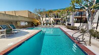 Located at 23825 Anza Ave in Torrance, California, this outdoor recreational area serves as a central amenity for the residential community. The centerpiece is a generously sized swimming pool with clear, blue-green water, framed by classic brick coping that adds a timeless, well-maintained aesthetic. Adjacent to the main pool is a circular spa, offering residents a dedicated space for relaxation. The surrounding sundeck is spacious, providing ample room for lounge chairs and seating arrangements, including a shaded umbrella table area ideal for enjoying the Southern California sunshine. The pool area is enclosed by a black wrought iron fence, ensuring both security and an open, airy feel. Mature trees, including tall palms and leafy deciduous varieties, are planted throughout the perimeter, offering natural shade and a lush, green atmosphere. The multi-story residential building borders the pool area, featuring balconies that overlook the courtyard, and a green-trimmed roofline that gives the property a distinctive character. The overall ambiance is bright, inviting, and well-suited to the warm climate of Torrance, making this outdoor space a practical and attractive gathering area for residents year-round.
