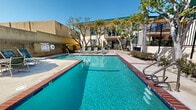 Located at 23825 Anza Ave in Torrance, California, this outdoor recreational area serves as a central amenity for the residential community. The centerpiece is a generously sized swimming pool with clear, blue-green water, framed by classic brick coping that adds a timeless, well-maintained aesthetic. Adjacent to the main pool is a circular spa, offering residents a dedicated space for relaxation. The surrounding sundeck is spacious, providing ample room for lounge chairs and seating arrangements, including a shaded umbrella table area ideal for enjoying the Southern California sunshine. The pool area is enclosed by a black wrought iron fence, ensuring both security and an open, airy feel. Mature trees, including tall palms and leafy deciduous varieties, are planted throughout the perimeter, offering natural shade and a lush, green atmosphere. The multi-story residential building borders the pool area, featuring balconies that overlook the courtyard, and a green-trimmed roofline that gives the property a distinctive character. The overall ambiance is bright, inviting, and well-suited to the warm climate of Torrance, making this outdoor space a practical and attractive gathering area for residents year-round.